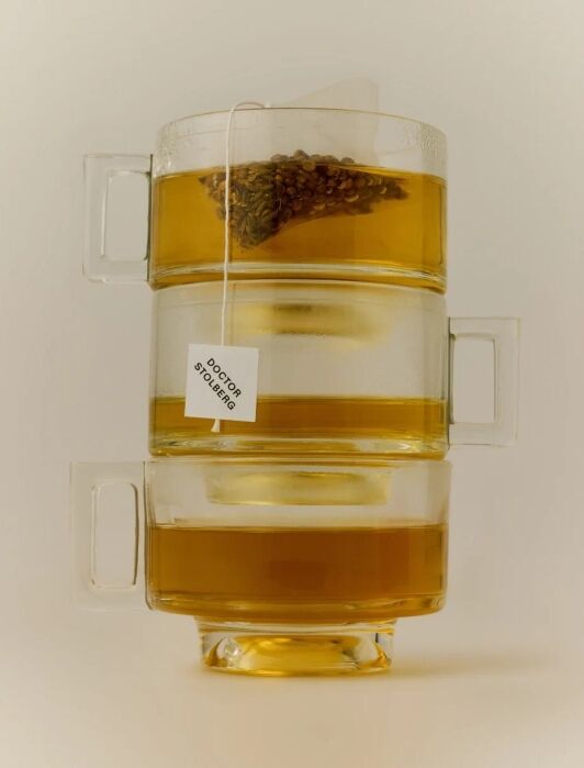 Three glass mugs stacked vertically, each filled with amber tea, with a tea bag labeled “Doctor Stolberg” hanging from the middle mug.