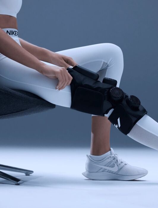 A person in white athletic wear adjusting a black therapeutic knee wrap with two circular control dials on their leg.