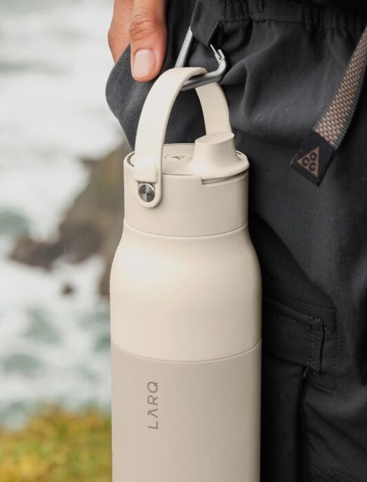 A beige LARQ water bottle with a handle hanging from a carabiner on a person’s pants.