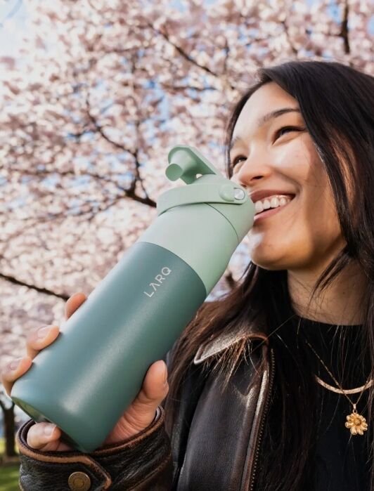 A smiling woman holds a two-toned sage green LARQ water bottle in front of a blooming cherry tree.