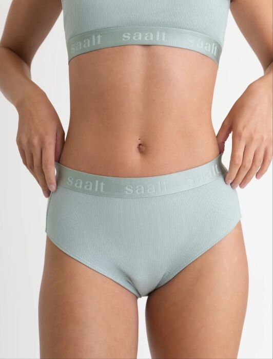 A person wearing a ribbed light green matching bralette and high-waisted underwear set with the brand name “saalt” printed on the waistbands.