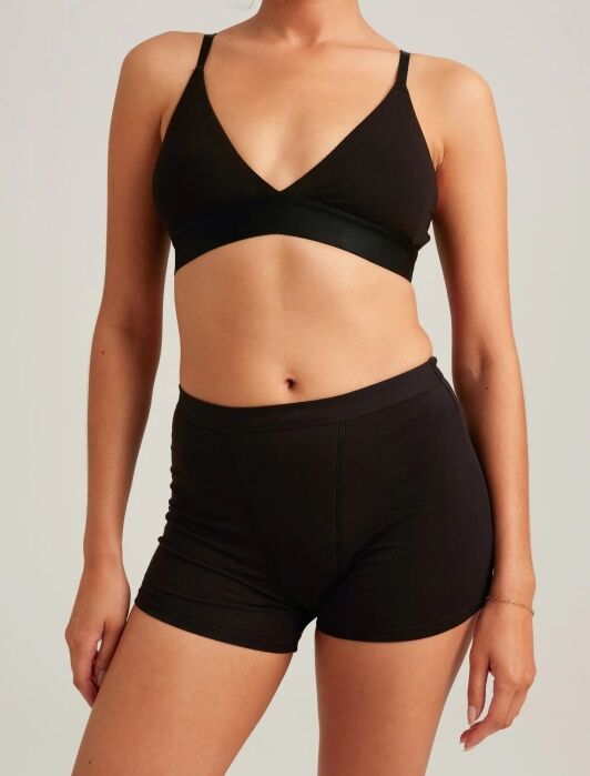 A person wearing a black bralette and black high-waisted shorts stands against a plain background, with their arms relaxed at their sides.