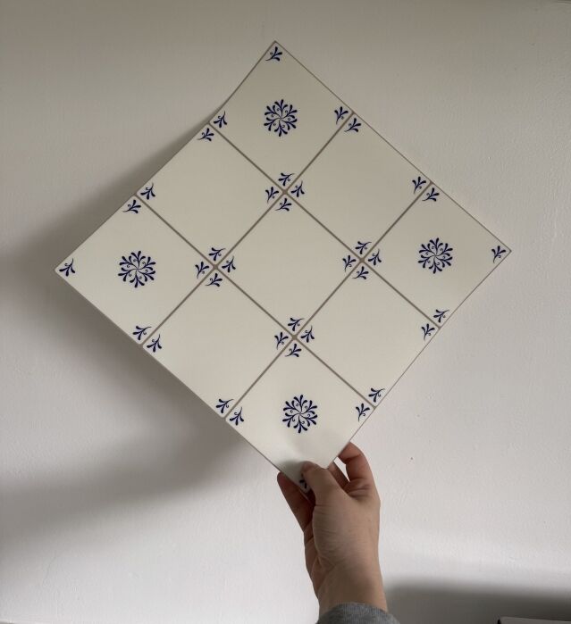 A hand holds a large ceramic tile composed of nine smaller white tiles with blue floral patterns, arranged in a 3x3 grid.