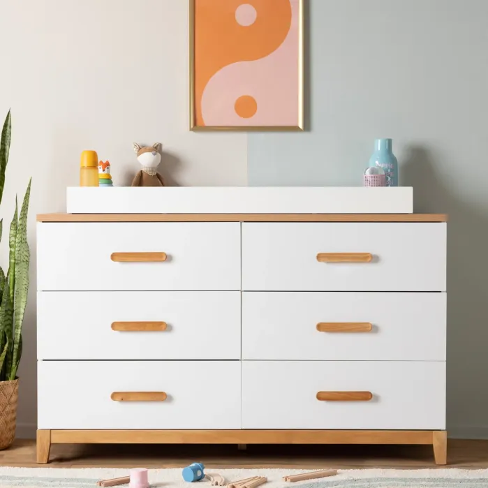 A white six-drawer dresser with wooden handles and a changing pad on top, featuring a framed abstract print on the wall above.