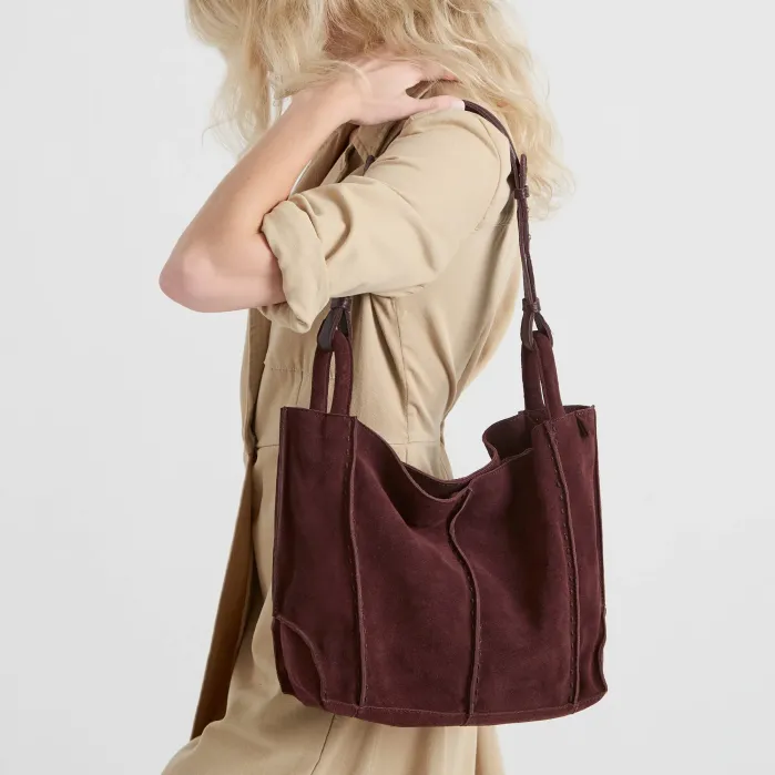 A person in a beige dress carrying a deep burgundy suede shoulder bag.