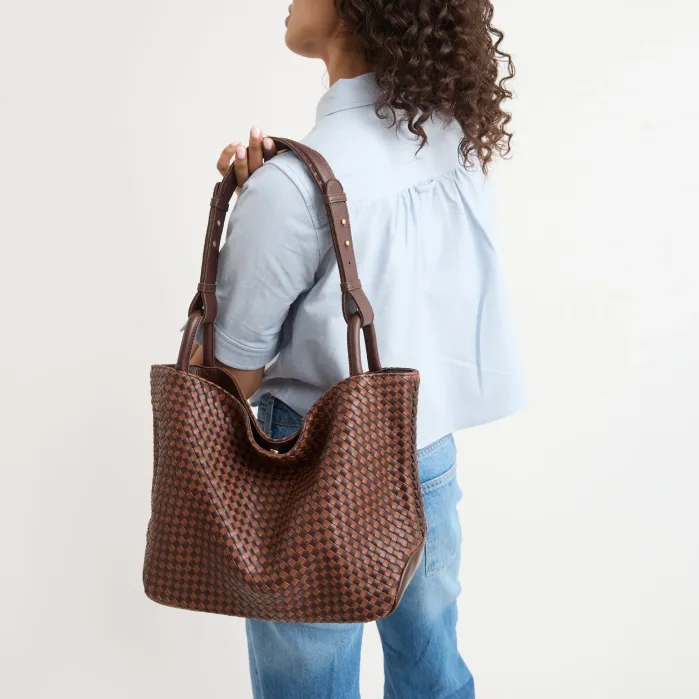 A person wearing a light blue shirt and jeans carries a large, dark brown woven leather tote bag over their shoulder.