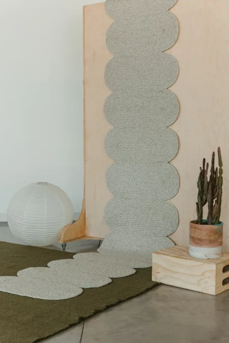 A light grey, scalloped runner rug mounted vertically on a wooden panel and extending onto the floor, next to a round paper lamp and a potted cactus.