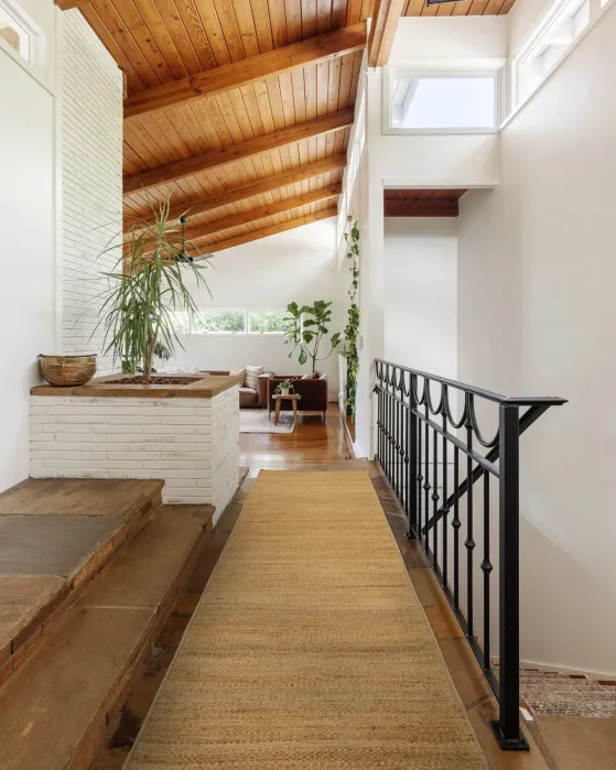 A view from a hallway looking into a living room with a vaulted wood ceiling, a white brick fireplace, and a black metal railing along the stairs.