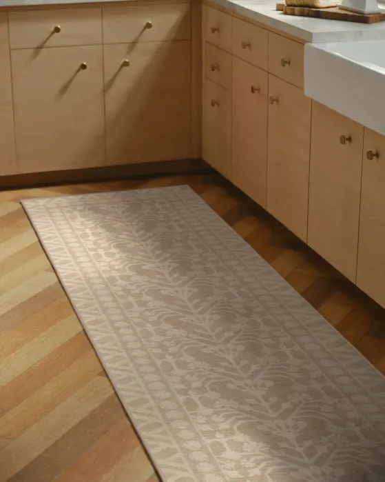 A beige runner rug with a repeating floral pattern placed on a herringbone wood floor in a kitchen with light wood cabinets.