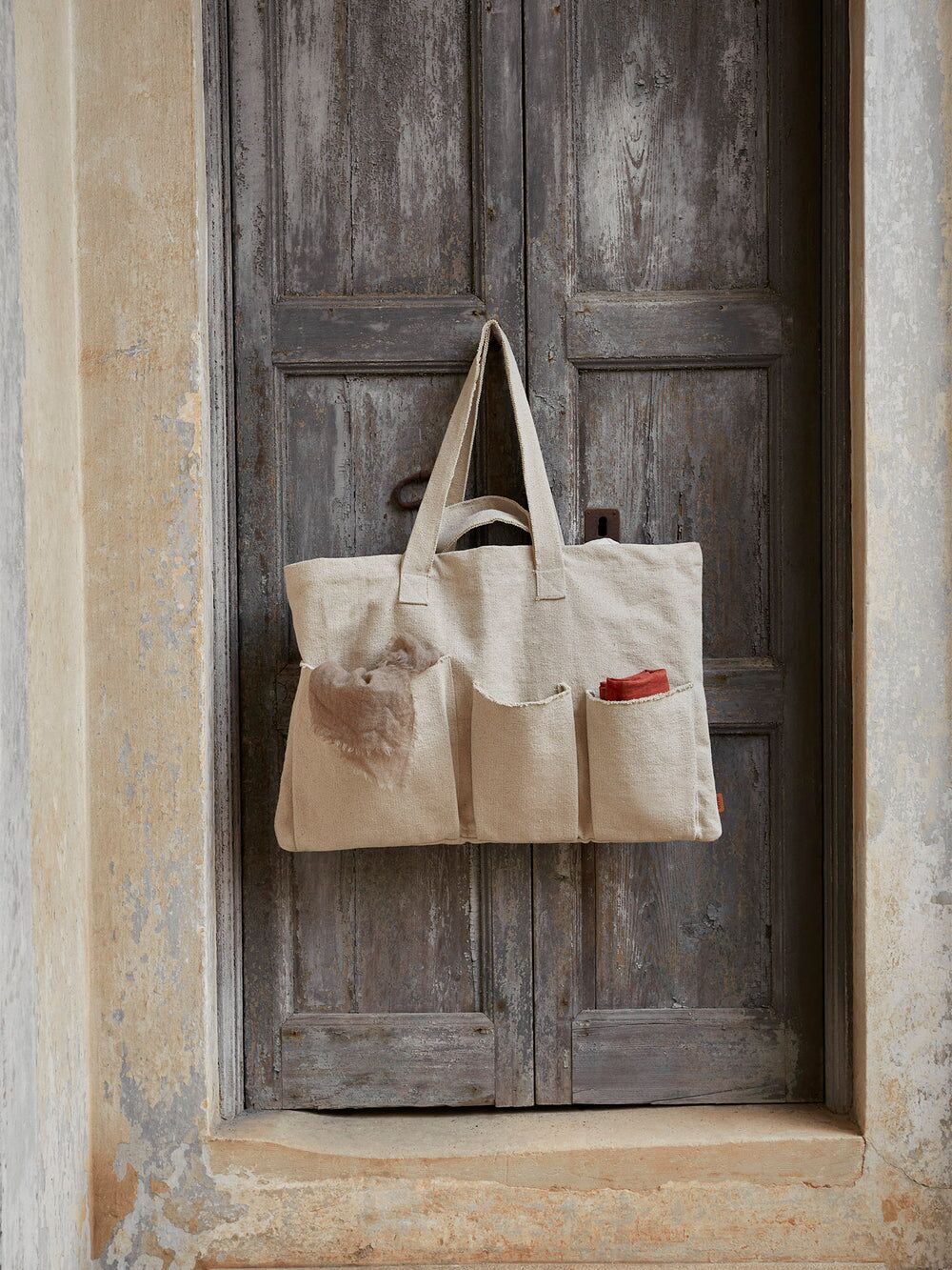Beige canvas tote bag with exterior pockets, hanging on a wooden door.