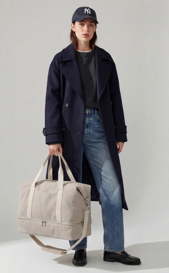 A person wearing a navy coat, blue jeans, and a New York Yankees baseball cap, holding a large beige duffel bag.