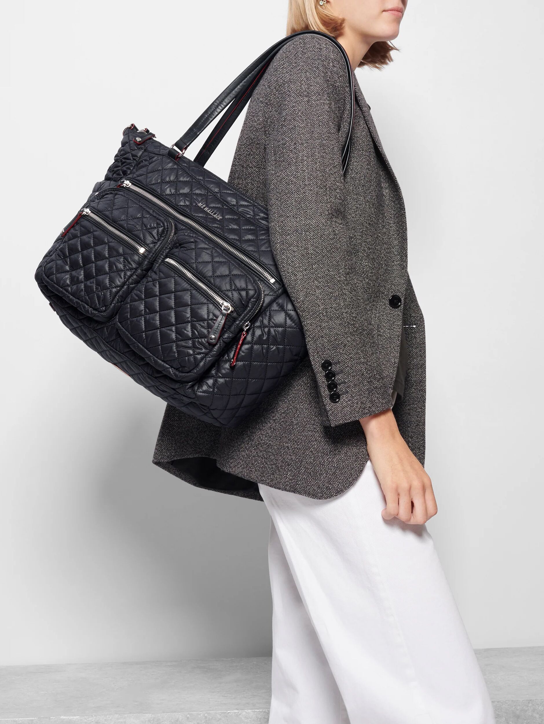 Person wearing a grey blazer and white trousers carrying a black quilted shoulder bag with multiple zippered pockets.