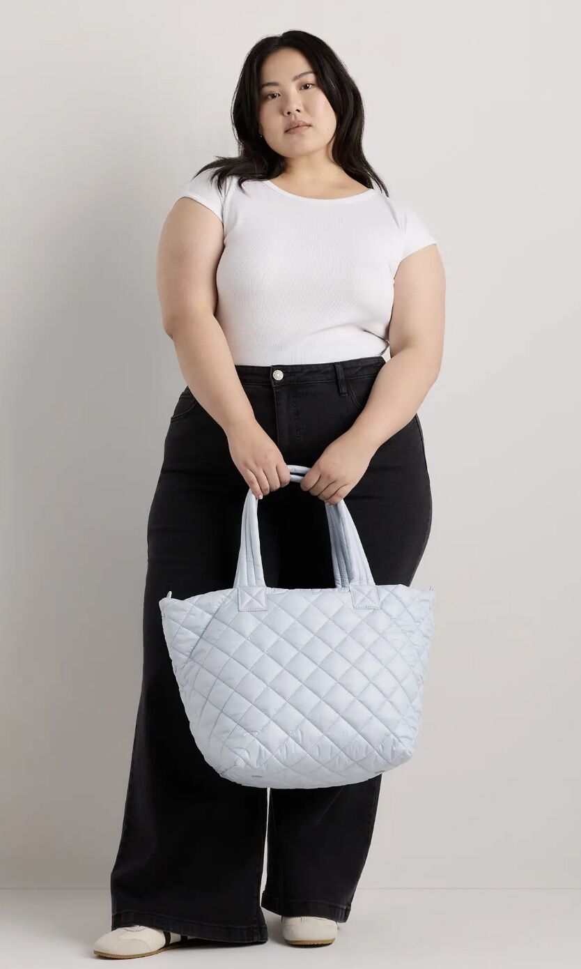 A person in a white t-shirt and black jeans holding a light blue quilted tote bag.