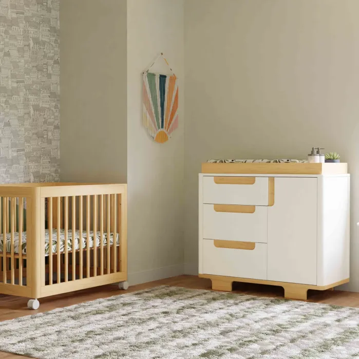 A wooden crib and a white dresser with three drawers, positioned on a textured rug in a nursery.