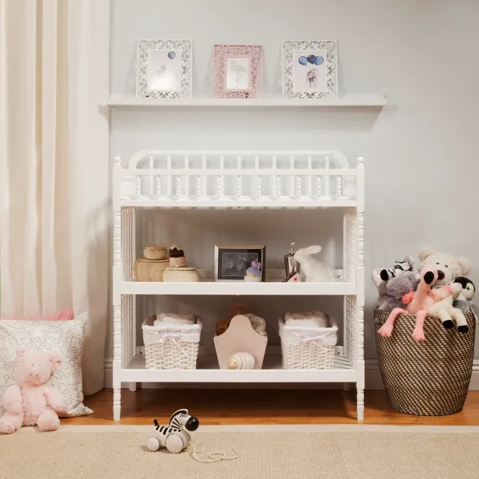 A white wooden changing table with shelves containing baskets, toys, and decor, flanked by a wicker basket of stuffed animals and a toy zebra on the floor.