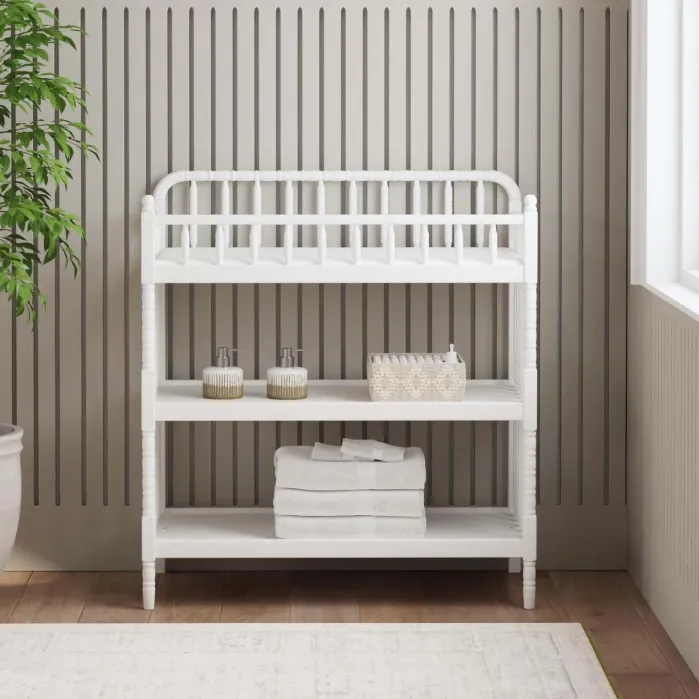 White wooden changing table with two open shelves holding two soap dispensers, a small basket, and stacked towels.