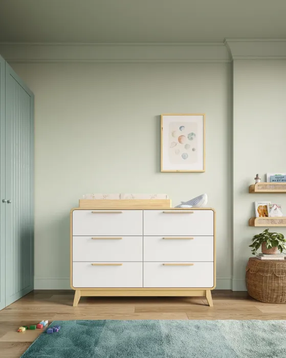 White six-drawer dresser with wooden trim and a changing pad on top, positioned in a nursery with a framed planet print on the wall.