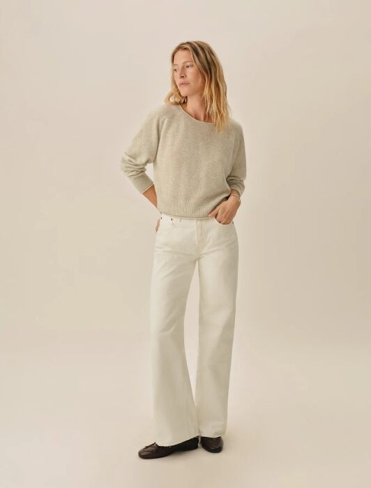 A person stands against a plain background wearing a light gray sweater, white wide-leg pants, and dark shoes, with hands in pockets and looking to the side.