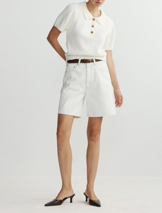 A person wears a short-sleeve white knit top with buttons, white high-waisted shorts, a brown belt, and black low-heeled shoes, standing against a plain light background.