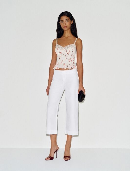 A woman stands against a plain white background wearing a floral camisole top, white cropped pants, maroon heels, and holding a black clutch.