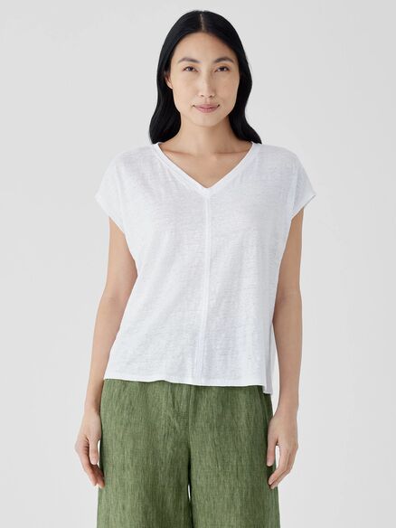 A person with long dark hair stands against a plain background, wearing a white V-neck short-sleeve shirt and green pants.