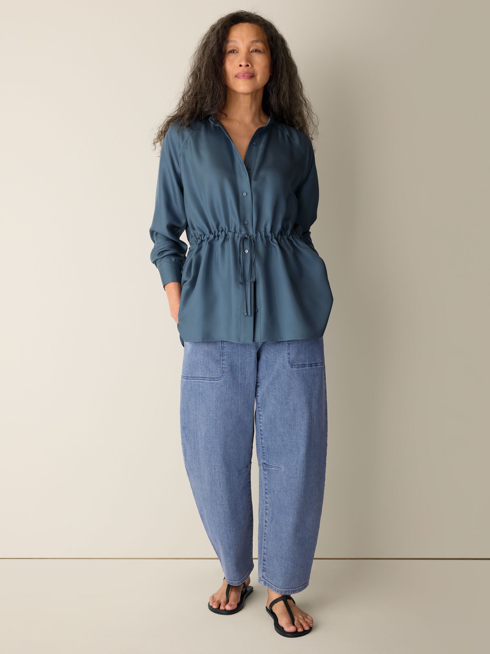A woman stands against a plain background wearing a teal button-up blouse with a drawstring waist, loose light blue jeans with front pockets, and black sandals.