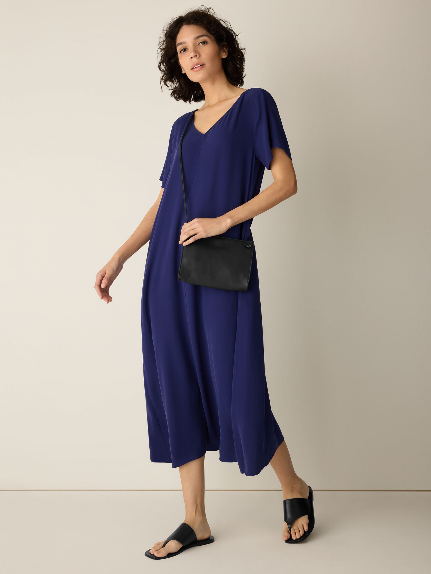 Woman wearing a loose-fitting, short-sleeve blue dress, black slide sandals, and a black crossbody bag, standing against a plain beige background.