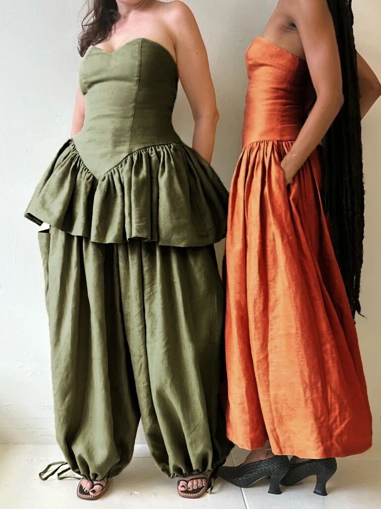 Two people wearing strapless, structured outfits; one in olive green with peplum and wide pants, the other in rust orange with a fitted bodice and full skirt; both standing against a plain wall.