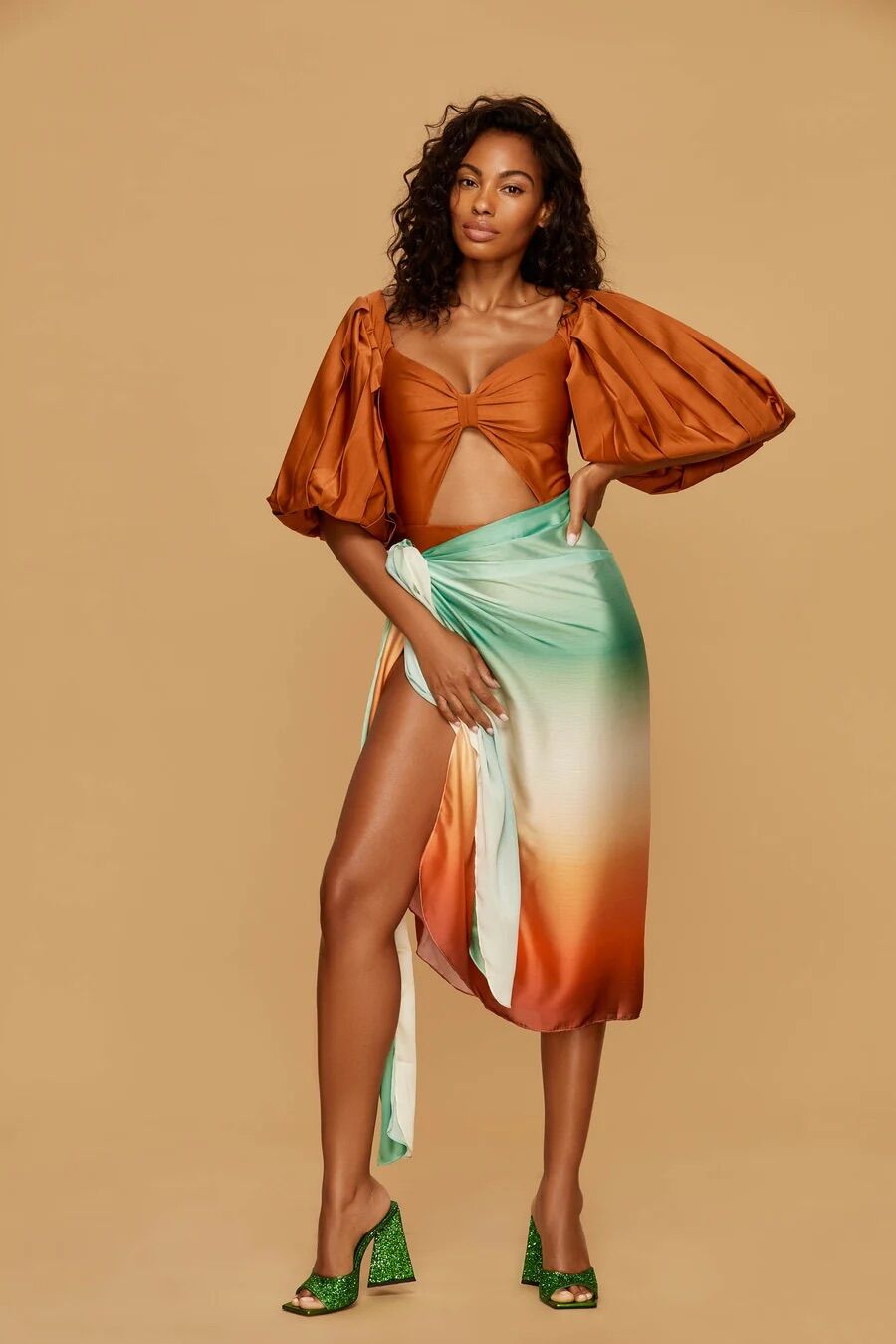 Woman stands against a tan background wearing a brown puff-sleeve top, a gradient green-orange wrap skirt, and green heeled sandals.