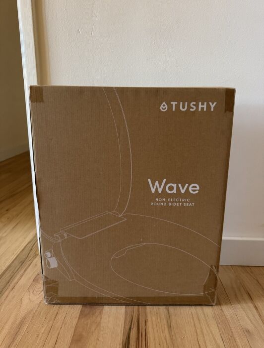 A boxed Tushy Wave non-electric round bidet seat rests upright on a wooden floor against a white wall.