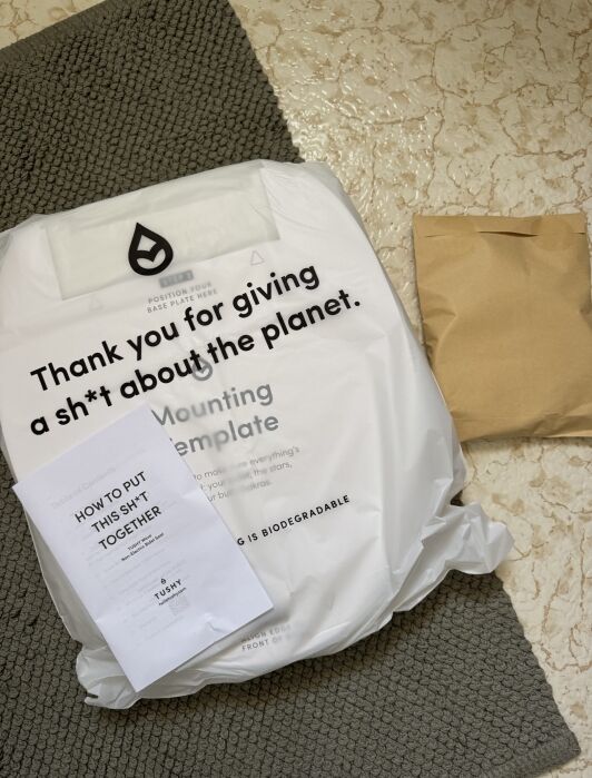 Biodegradable package on a bath mat with a paper guide titled “HOW TO PUT THIS SH*T TOGETHER” and a brown envelope, all on a tiled floor.