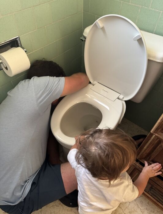 An adult is fixing a toilet while a young child watches and touches the toilet seat in a bathroom with green tiles.