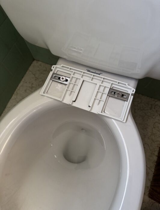 A toilet with its seat removed, exposing the mounting bracket and screws on the rim, in a bathroom with green tile walls and a beige patterned floor.
