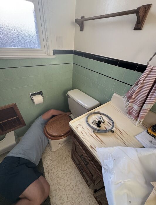 A person is lying on the bathroom floor next to a toilet, appearing to make repairs. The toilet seat is removed and placed on the sink, along with some tools.