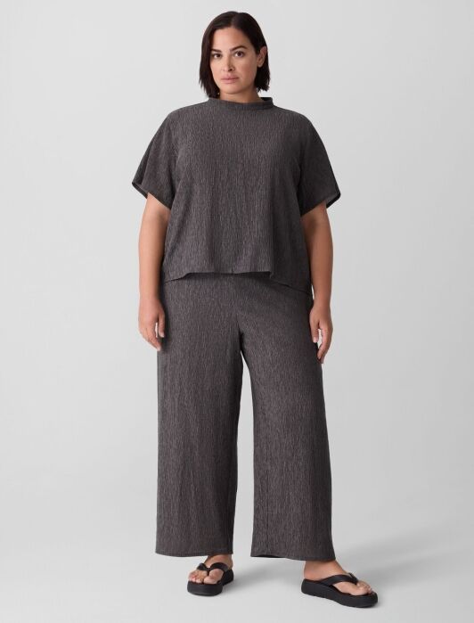A person stands facing forward, wearing a loose-fitting, dark gray textured top and matching wide-leg pants with black sandals, against a plain light background.