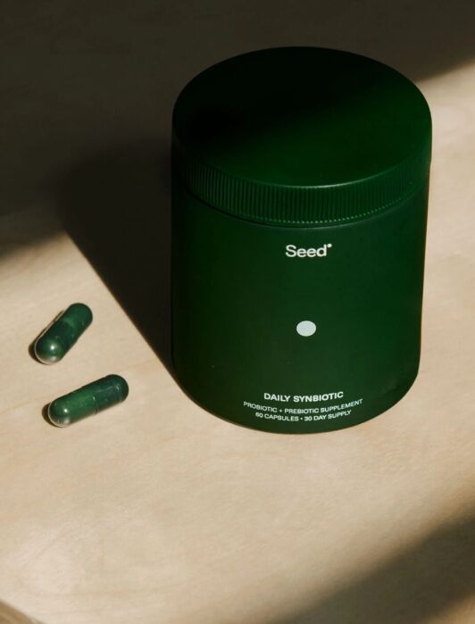 A jar of Seed DS-01 Daily Synbiotic next to two green gel cap pills in a spotlight.