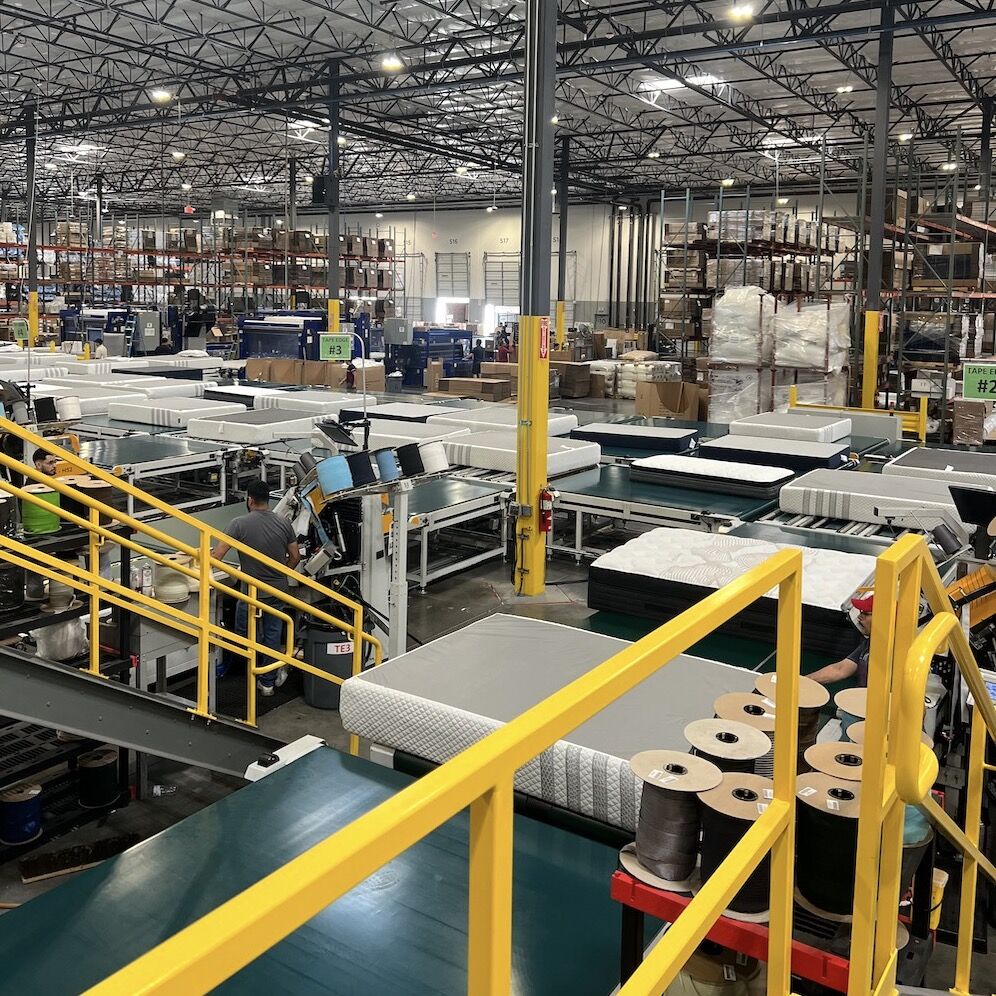 A large industrial factory interior buzzes with the energy of workers operating machinery and conveyor belts. Amidst the hum, shelves stocked with nontoxic mattress materials stand in the background, supporting production activities while ensuring a safe and eco-friendly environment.