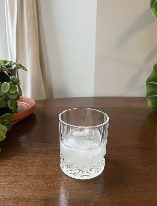 A clear glass with ice and a light-colored beverage sits on a wooden table next to a potted plant.