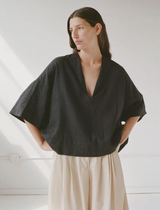 Woman in a black blouse and beige pants standing in a sunlit room, hands on hips, looking to the side.