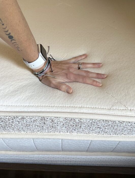 Reviewer Randi Donahue's hand pressing down on an organic Birch mattress, demonstrating its texture and responsiveness. The person is wearing a smartwatch and several bracelets, making it clear this isn't just a mattress review but a glimpse into lifestyle choices aligned with comfort and sustainability.