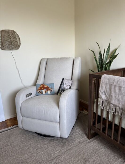 A light gray recliner sits next to a wooden crib with a beige blanket. Two children's books rest on the chair, and a green plant is on the floor beside it.