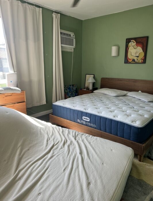Bedroom with green walls featuring two beds, one with a blue mattress labeled "Elite Hybrid" and the other bare. The room has a wall-mounted air conditioner, white curtains, and a framed painting.