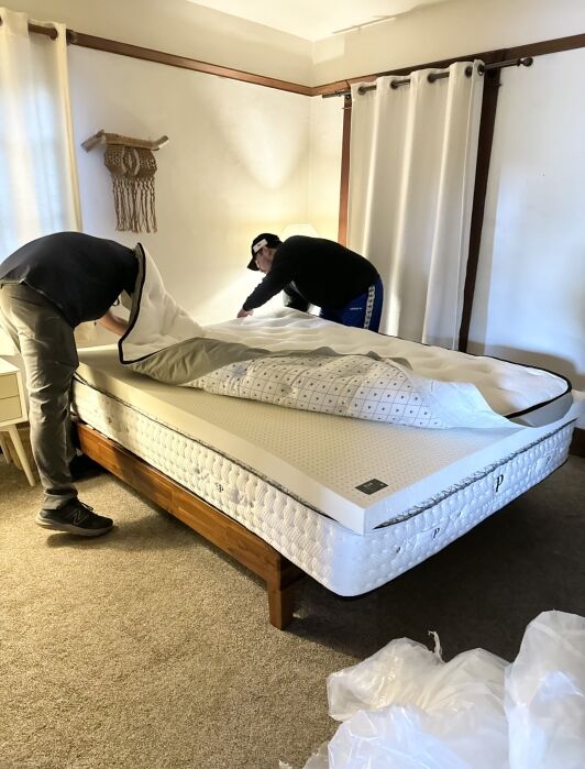 Two people are making a bed with a white mattress on a wooden frame, adjusting a mattress cover in a well-lit room.