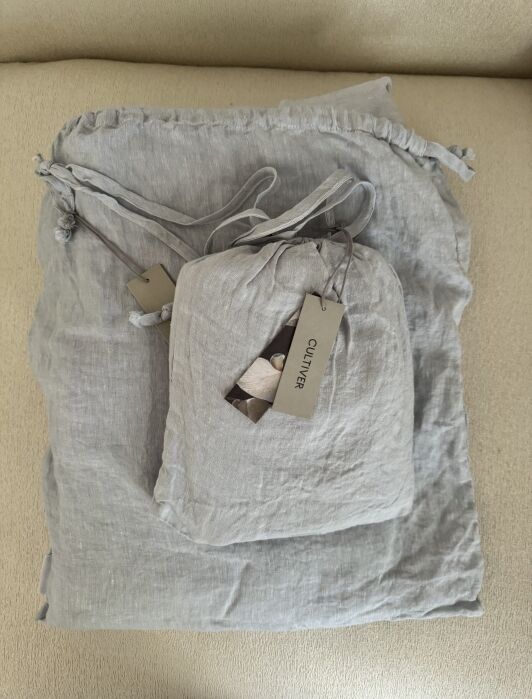 Two gray, drawstring linen bags with tags attached, placed on a light-colored surface.