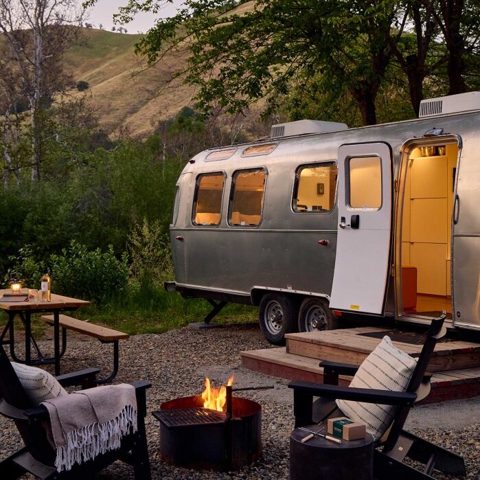 A silver camper trailer is parked at a campsite with a fire pit, patio chairs, a picnic table, and surrounding trees and hills in the background.