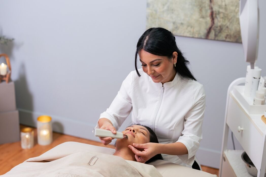 A skincare professional uses a handheld device on a client’s face during a facial treatment in a spa setting.