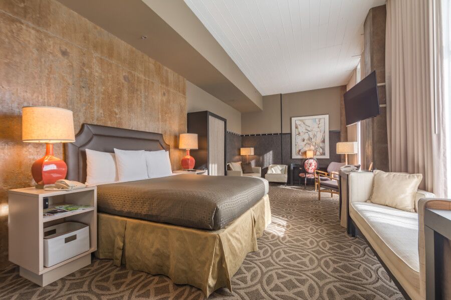 A hotel room with a king bed, two bedside tables with lamps, a sofa, a flat-screen TV, and large windows with curtains.