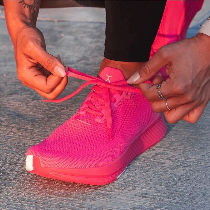 Person tying the laces of a bright pink running shoe on a concrete surface.