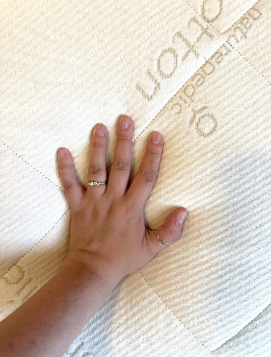 A hand with two rings presses down on a cream-colored, textured mattress with stitched patterns and partial text visible.