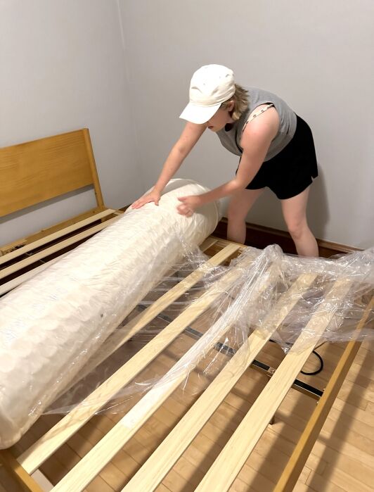 A person in a hat unrolls a vacuum-sealed mattress onto a wooden bed frame in a sparsely furnished room.